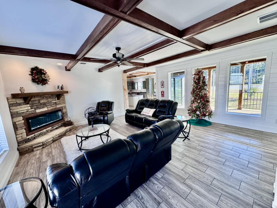 Spacious living room with black leather sofas, a stone fireplace, and a Christmas tree. Large windows bring in natural light, creating a cozy atmosphere.