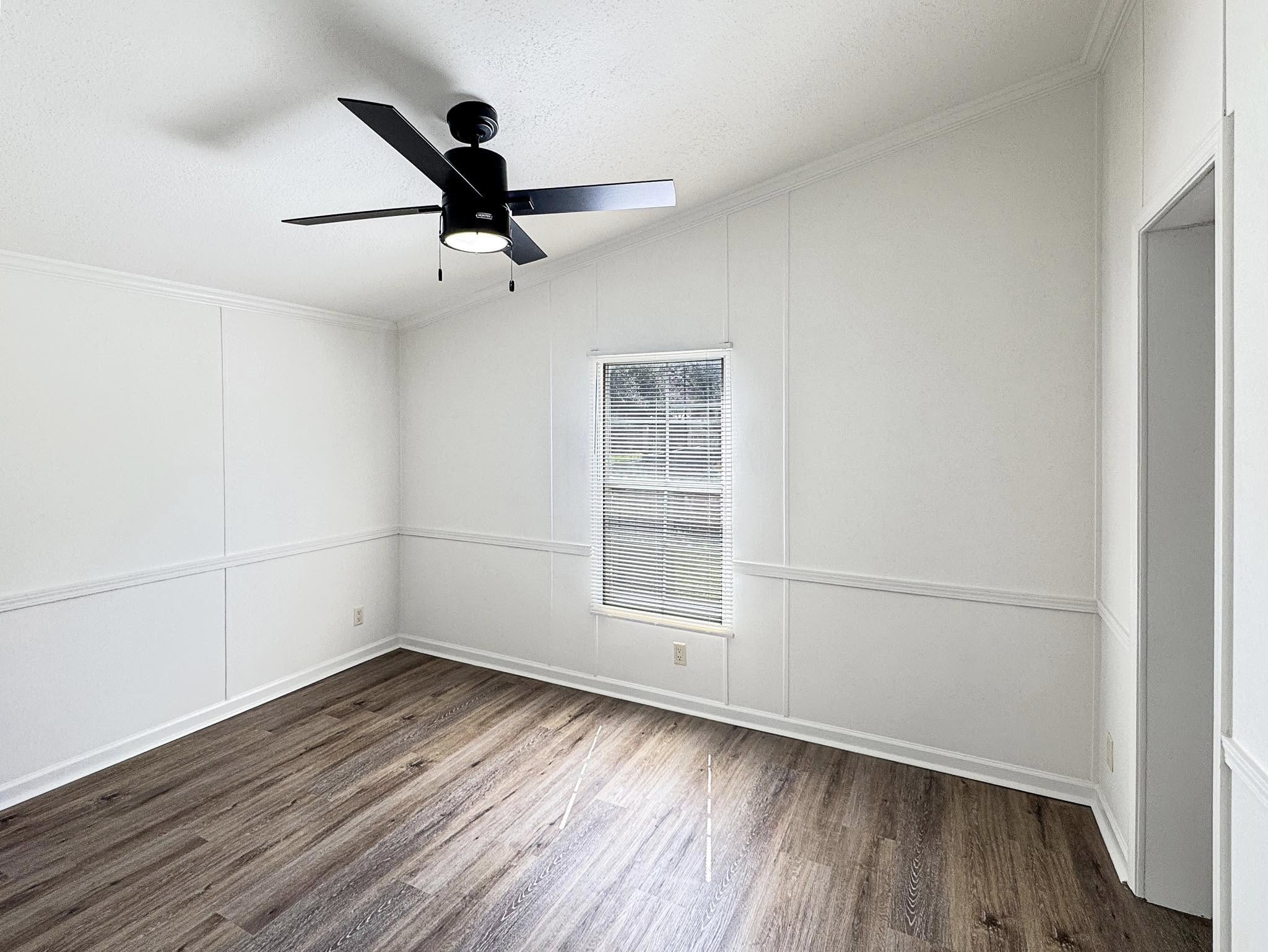A bright, empty room with white walls and a wood floor. A modern black ceiling fan hangs from the ceiling. Natural light enters through a window with blinds.