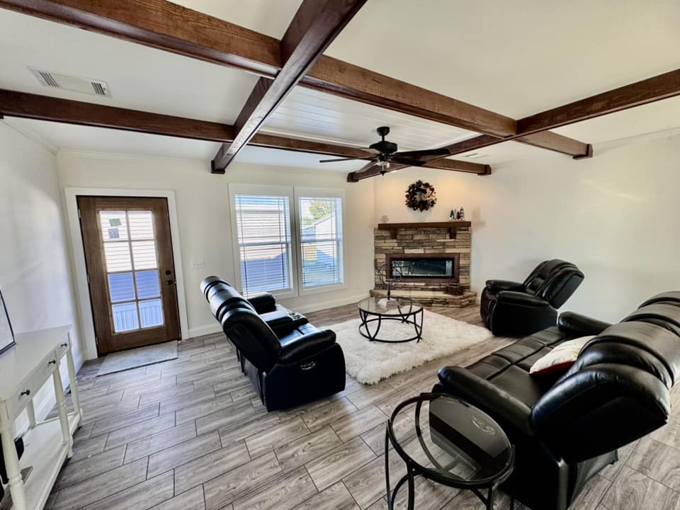 Spacious living room with black leather sofas, a stone fireplace, and a Christmas tree. Large windows bring in natural light, creating a cozy atmosphere.