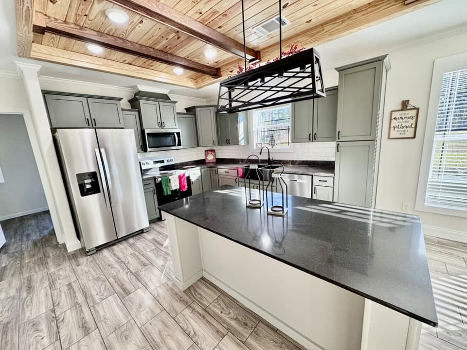 Spacious kitchen with gray cabinets, stainless steel appliances, and a black island counter. Wooden ceiling beams and modern lighting add warmth.