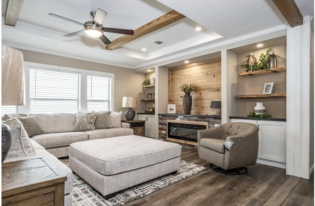 Cozy living room with beige sectional sofa, large ottoman, and swivel chair. Features wood accents, shelves with plants, modern fireplace, and ceiling fan.