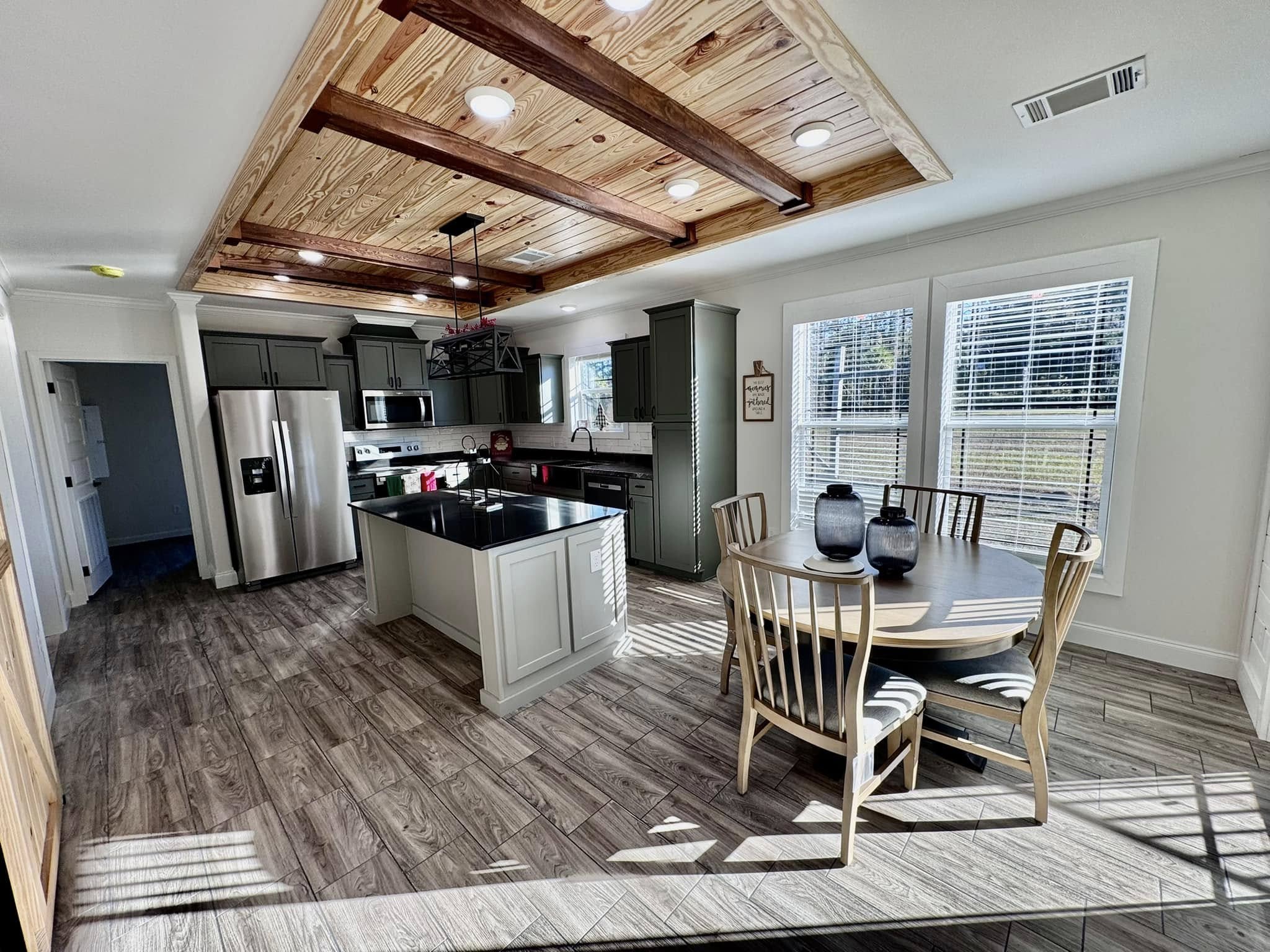 Modern kitchen with wooden ceiling beams, pendant lights, and gray cabinetry. A round dining table with chairs sits by sunlit windows, creating a warm, inviting atmosphere.