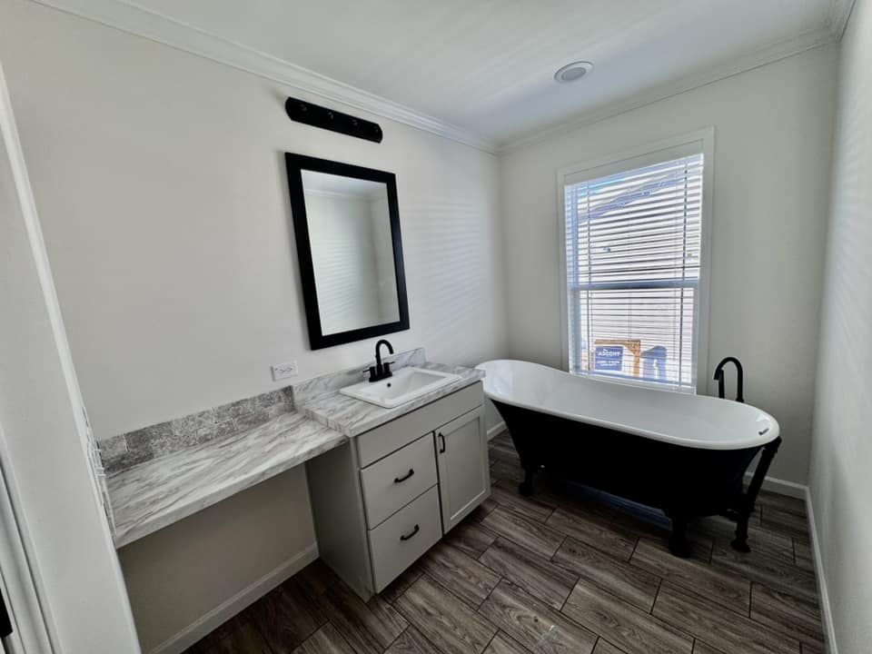 Modern bathroom with a large black freestanding tub next to a window with blinds. A marble countertop and sink with a black-framed mirror are visible, set against white walls and wood-style flooring.