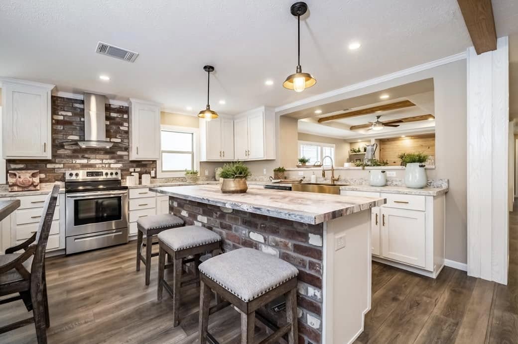 Modern kitchen with a large island, brick accents, and marble countertops. White cabinets, pendant lights, and wooden stools create a welcoming atmosphere.