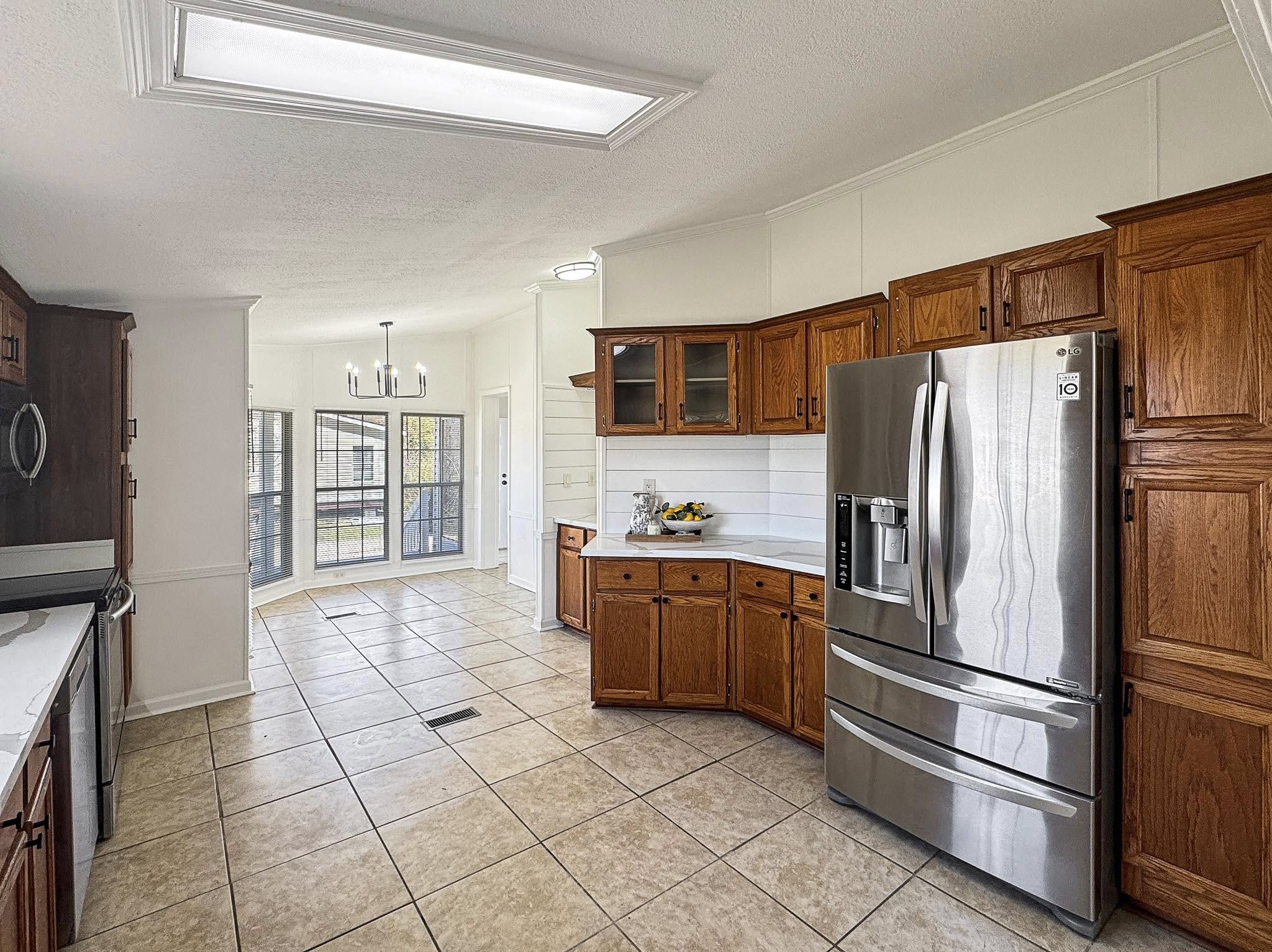 Spacious kitchen with wooden cabinets, stainless steel fridge, and beige tiled floor. Sunlit dining area with a modern chandelier and large windows.