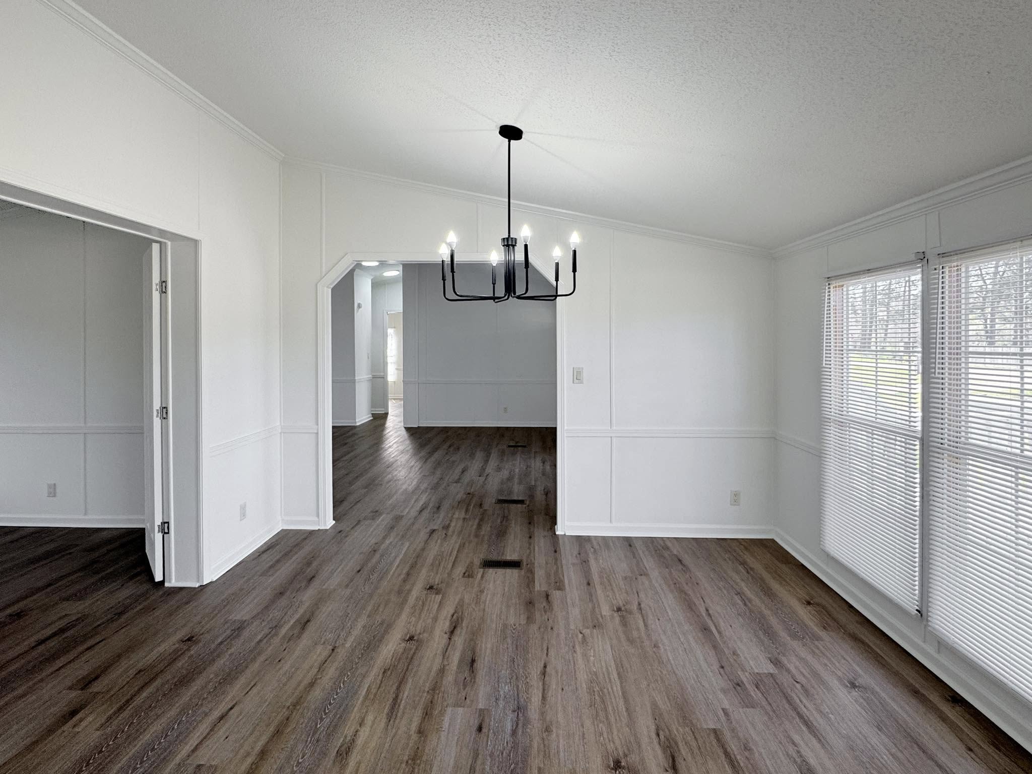 Spacious, bright room with white walls and wooden floors. Features a modern black chandelier and large windows with blinds, creating an airy atmosphere.