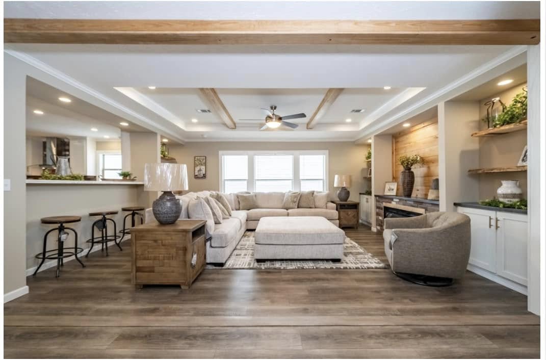 Spacious living room with light wood floors, beige sectional sofa, large ottoman, and wooden ceiling beams. Cozy seating, warm tones, inviting atmosphere.