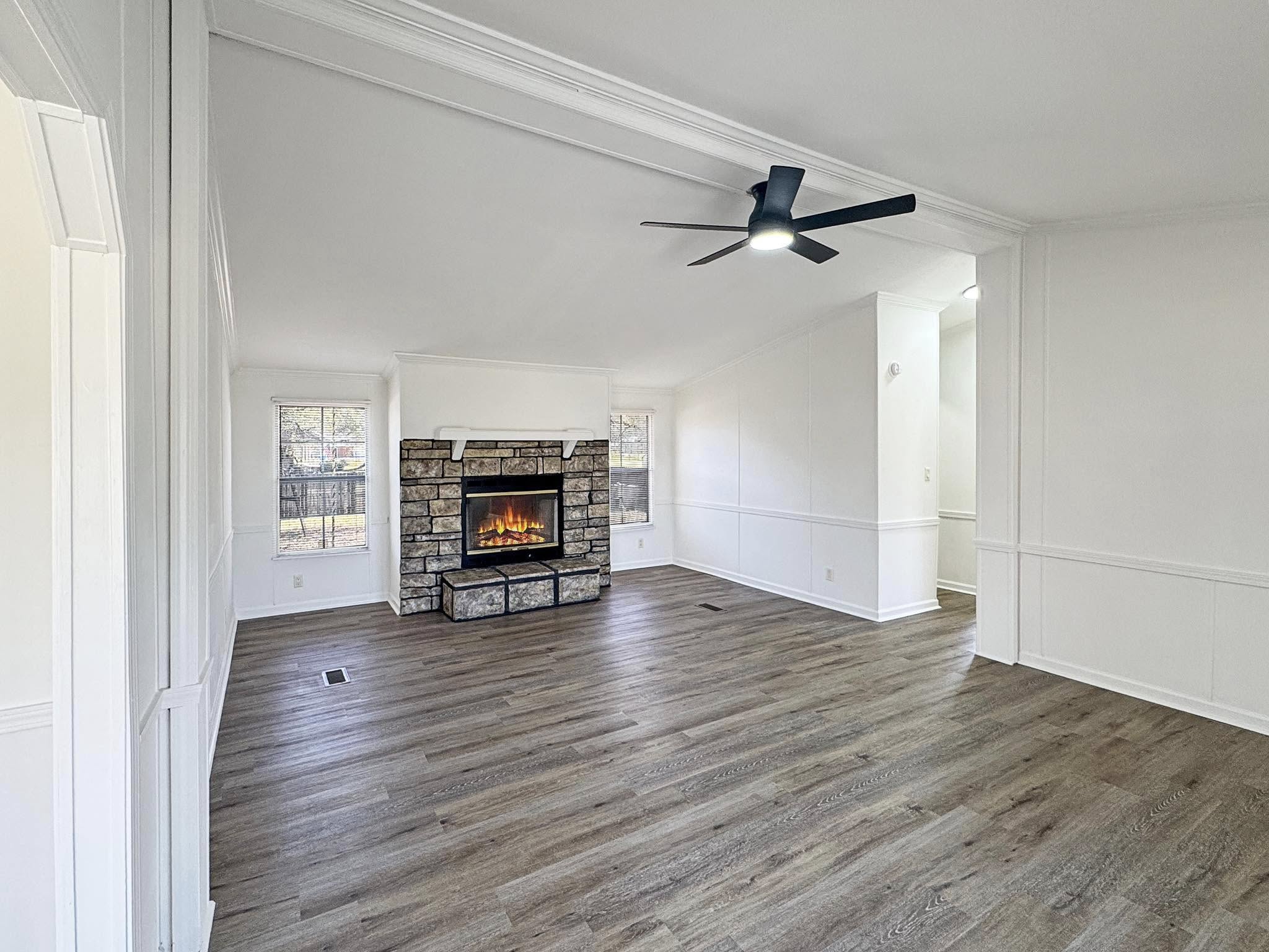 A spacious, light-filled living room features a rustic stone fireplace and modern ceiling fan. Large windows and wood floors add warmth and elegance.