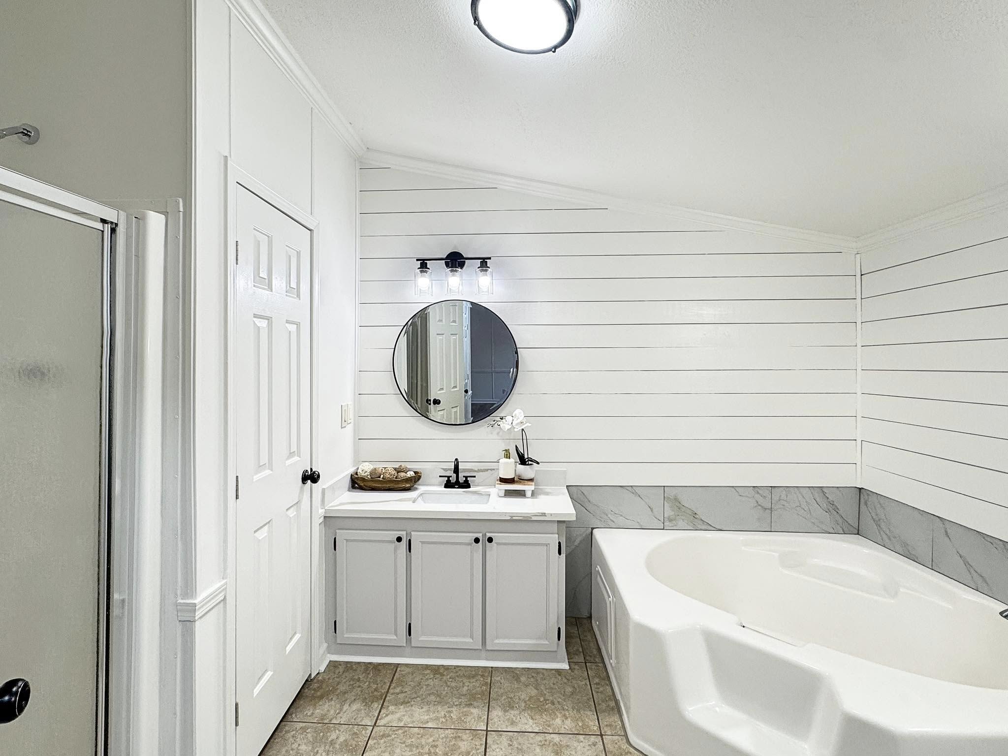 Modern bathroom with white shiplap walls, featuring a round mirror above a white vanity. A large corner bathtub is beside it, creating a serene atmosphere.