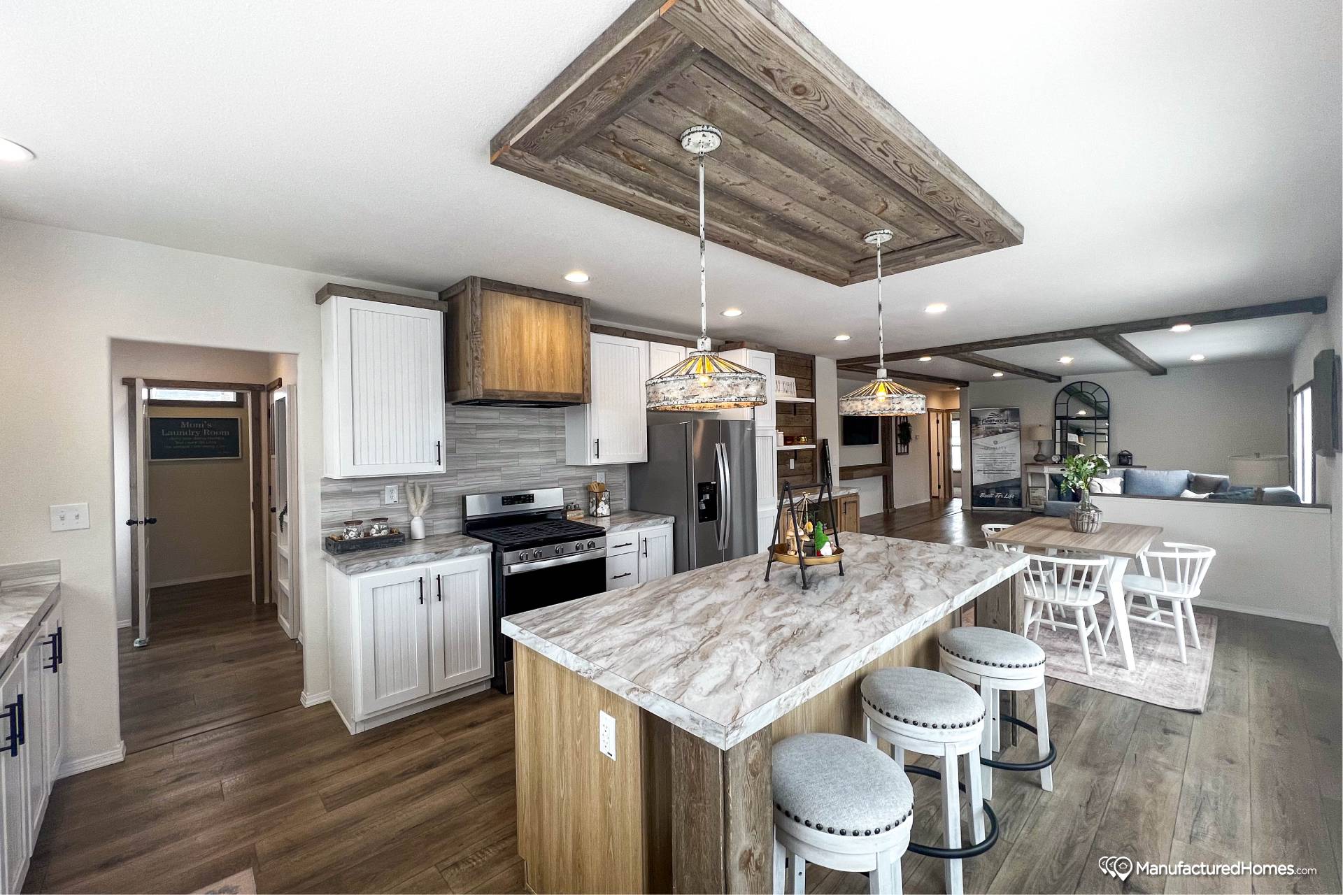 Modern kitchen with wooden floors, marble island, and three cushioned stools. White cabinets, stainless steel appliances, and pendant lights add elegance. Cozy, open layout leads to a dining area.