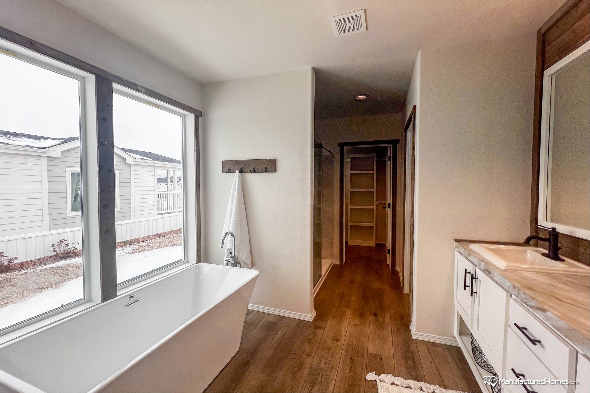 Modern bathroom with large freestanding bathtub by a window, wooden floor, white walls, double sink with sleek fixtures, and a walk-in closet.