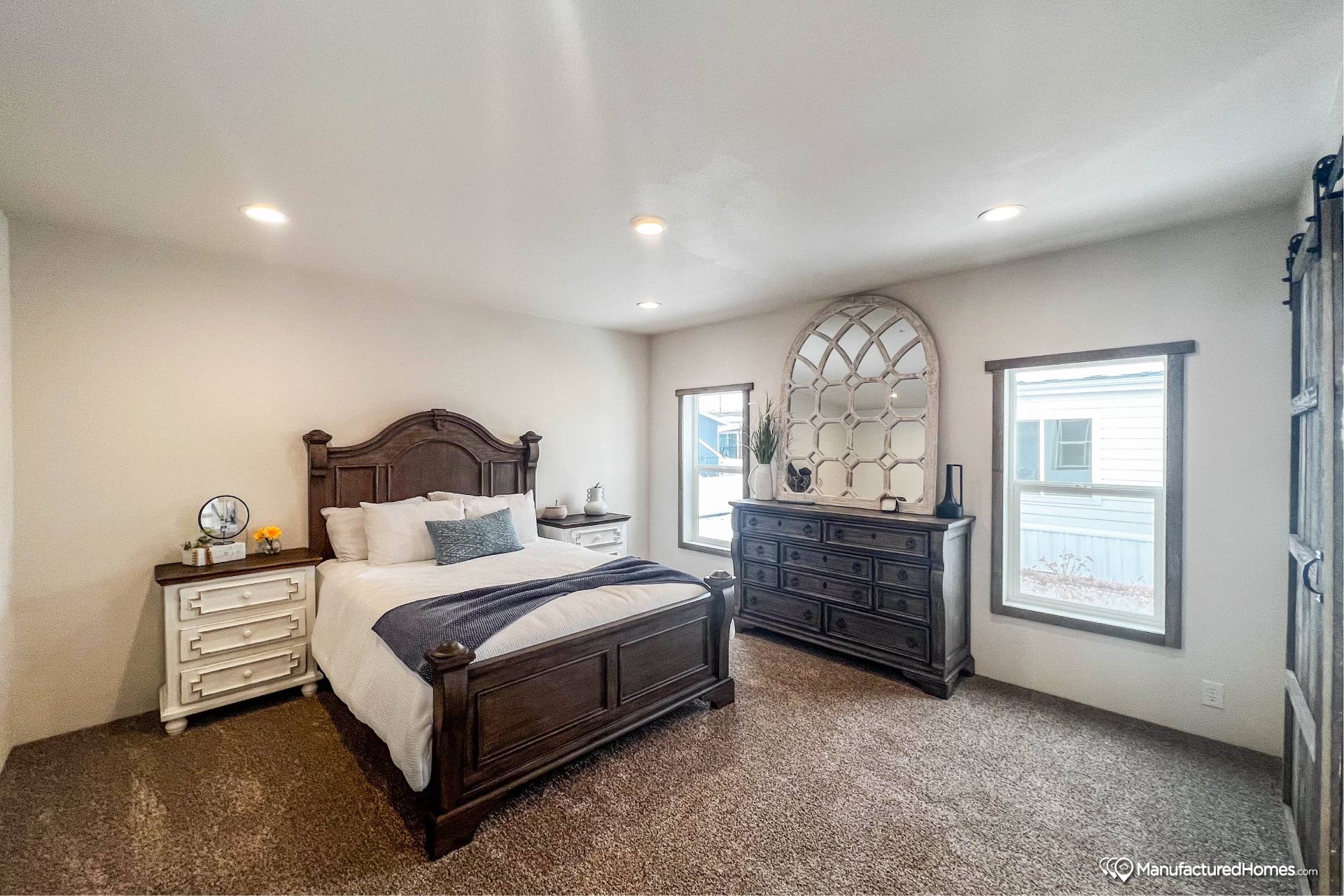 A cozy bedroom features a dark wood bed with white linens and a navy throw, flanked by white nightstands. A large dresser with a decorative mirror sits between two windows, showcasing a calm and inviting atmosphere.