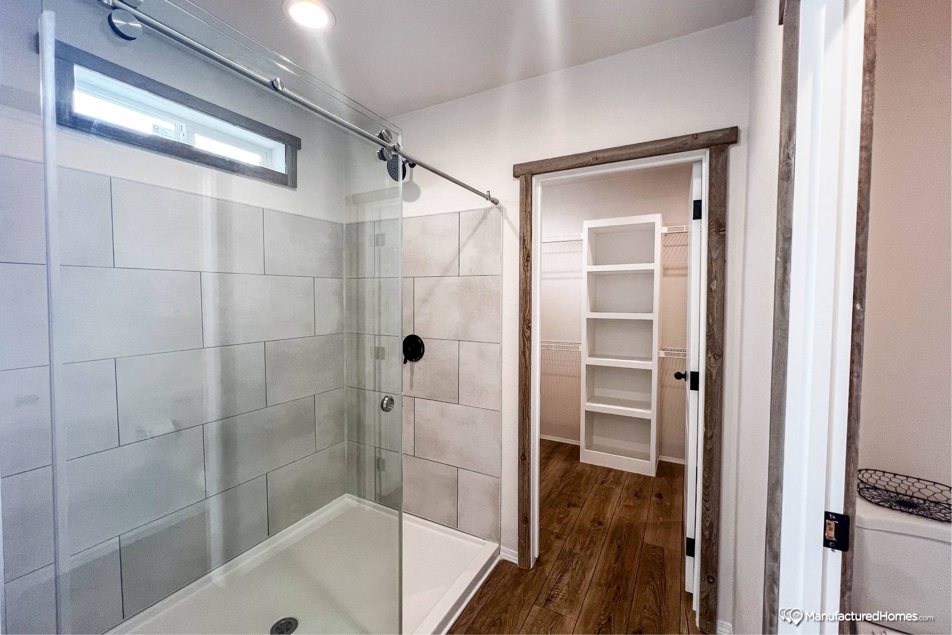 Modern bathroom with a glass-enclosed shower featuring gray tiles, a wood-floored walk-in closet in view. Bright and clean design with a serene feel.