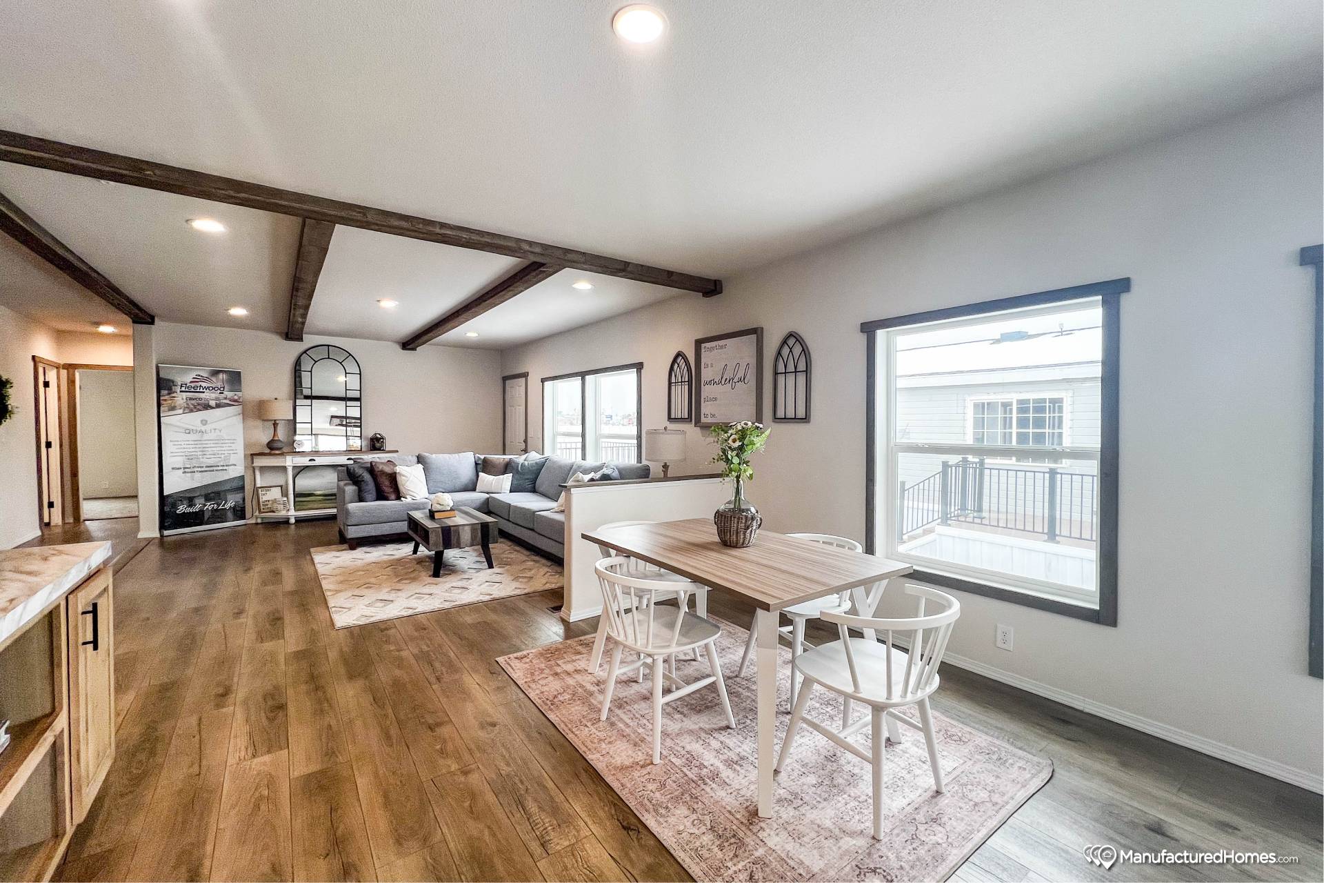 Spacious living and dining area with wooden floors and ceiling beams. Cozy gray sofa in living space; modern dining table with chairs in a bright room.