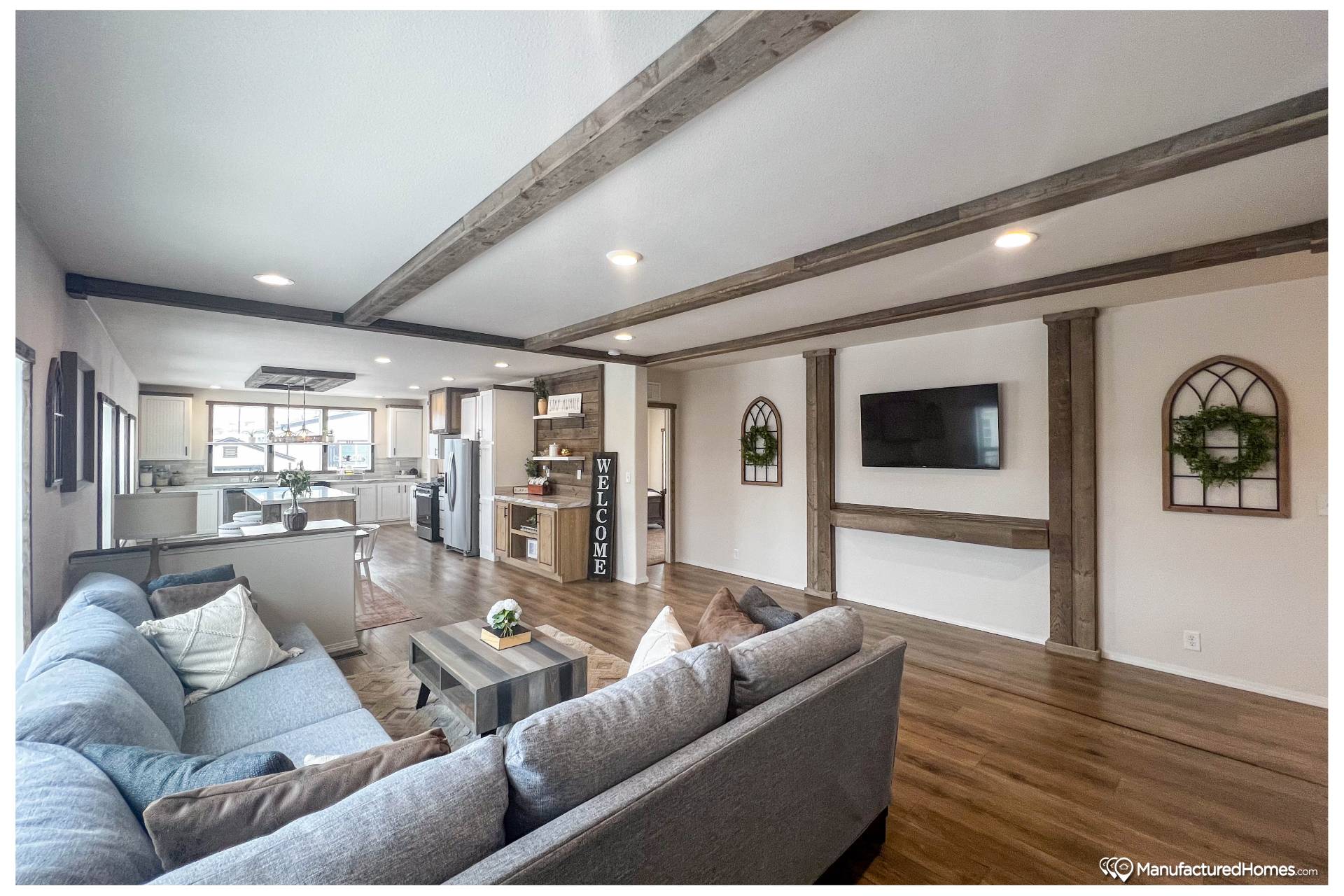 Spacious living room with a cozy gray sectional sofa, wooden ceiling beams, and a TV on a white wall. Open floor plan leads to a modern kitchen. Warm and inviting ambiance.
