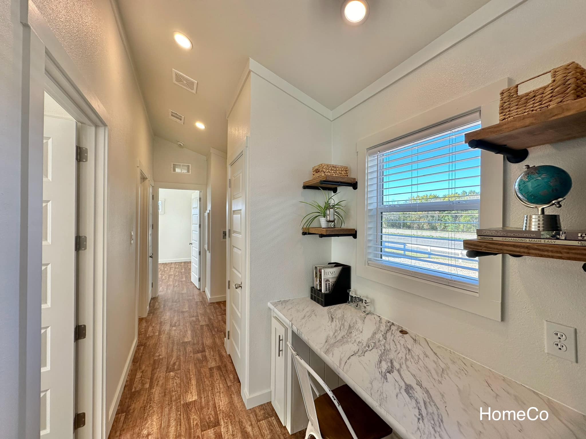 Bright, modern hallway with warm wood floors. White doors line the left wall. Right side features a small marble desk, shelves, and a window view. Cozy and inviting.