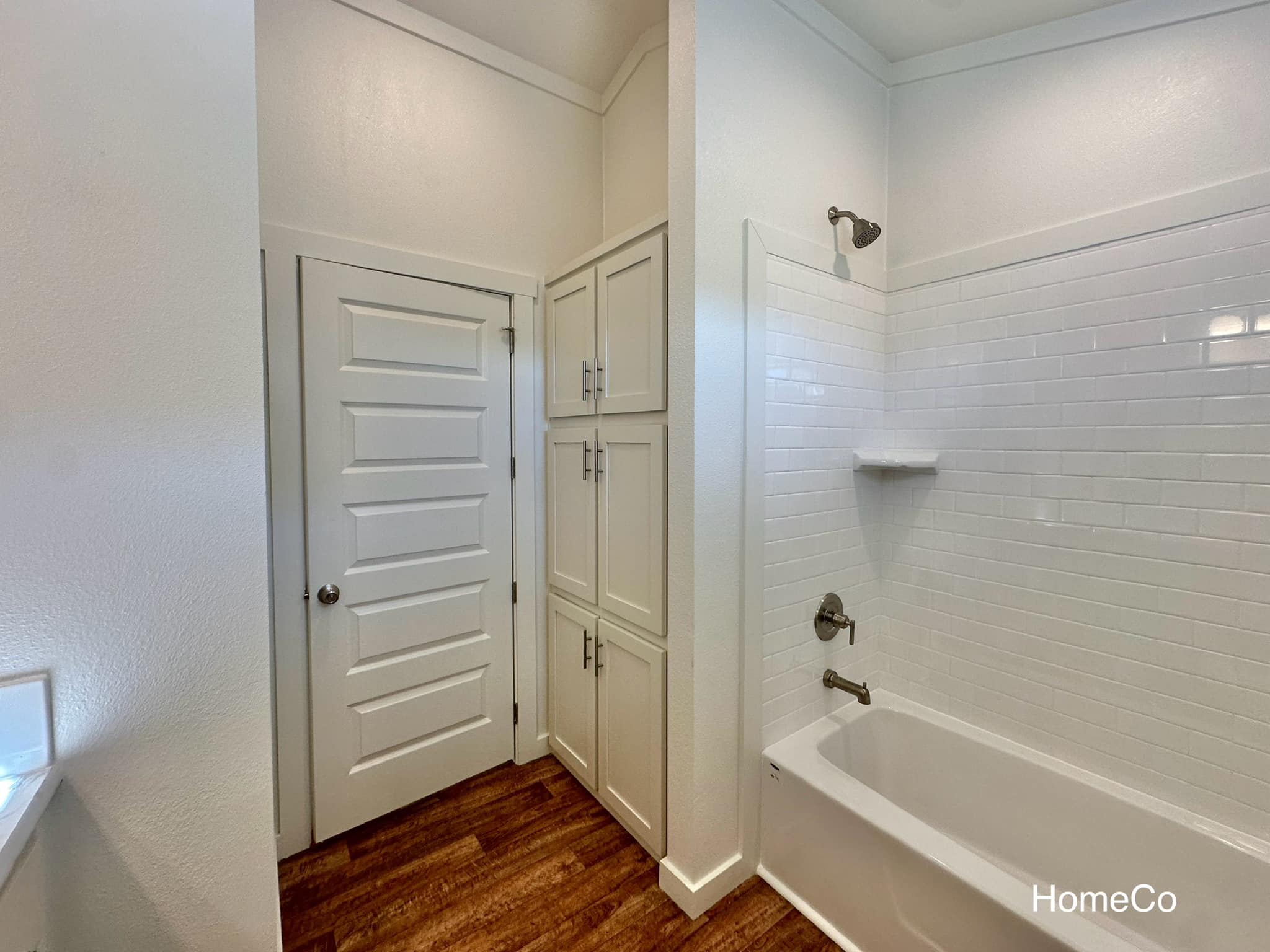 Bright, modern bathroom with white subway-tiled bathtub, wooden cabinets, and a paneled door. Warm wood floor adds a cozy, inviting touch.