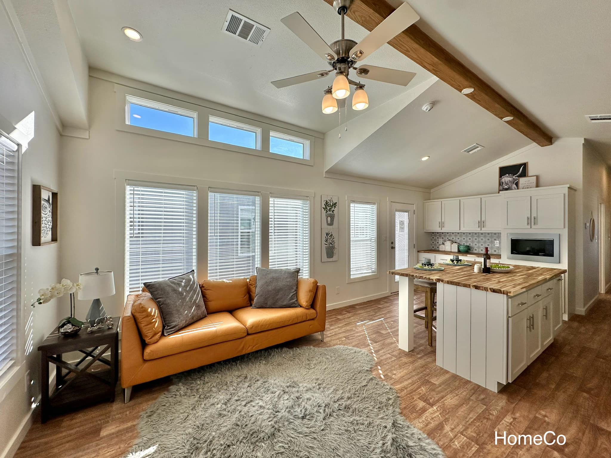 Bright, modern living area with high ceilings features a tan leather sofa, gray cushions, a fluffy rug, and a wooden-beam ceiling fan. The adjacent kitchen has a cozy island with stools, white cabinetry, and a warm ambiance.