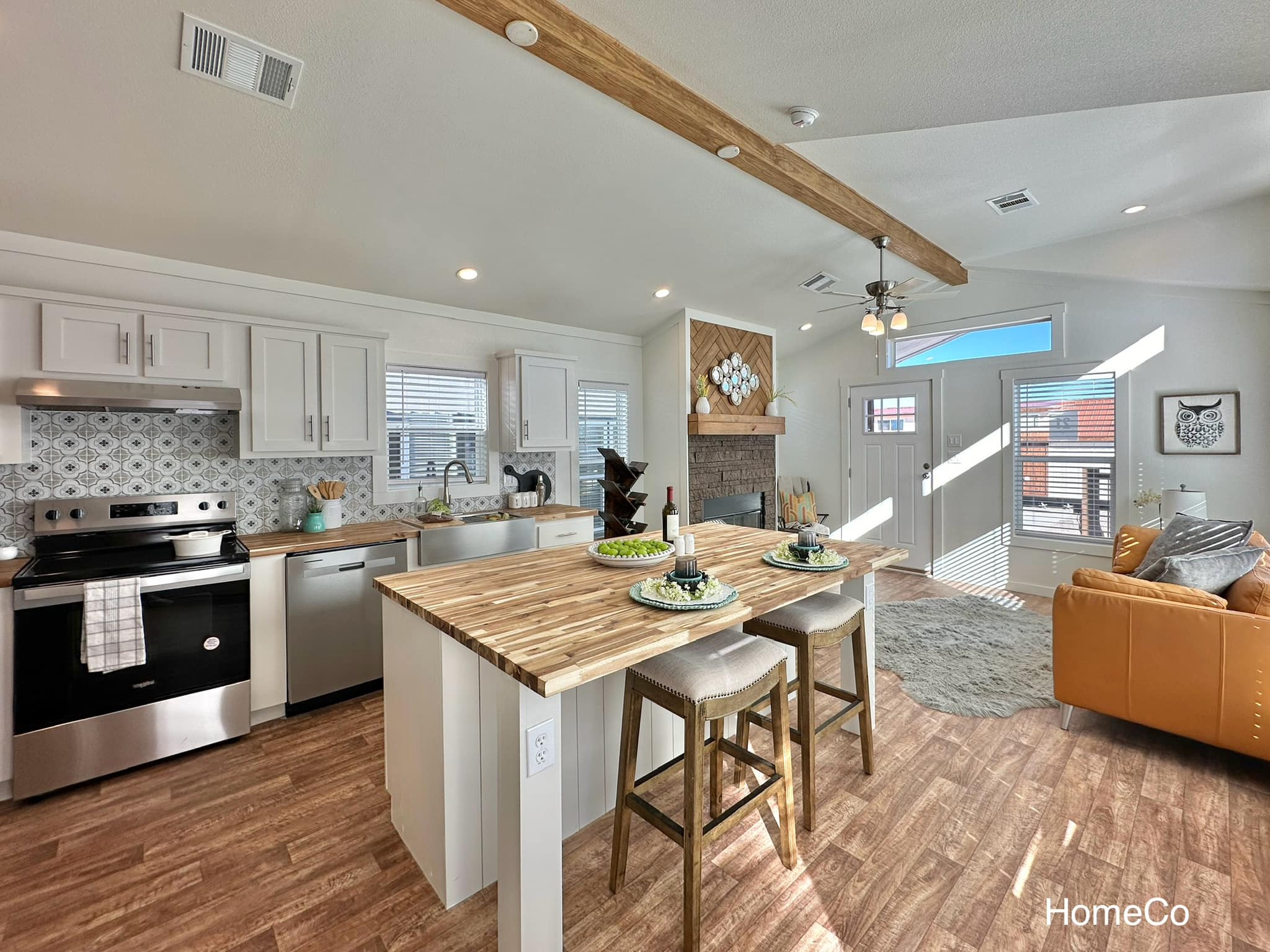 Modern kitchen and living area with a bright, airy feel. Features a wooden island with bar stools, stainless steel appliances, and a cozy sofa near a fireplace.