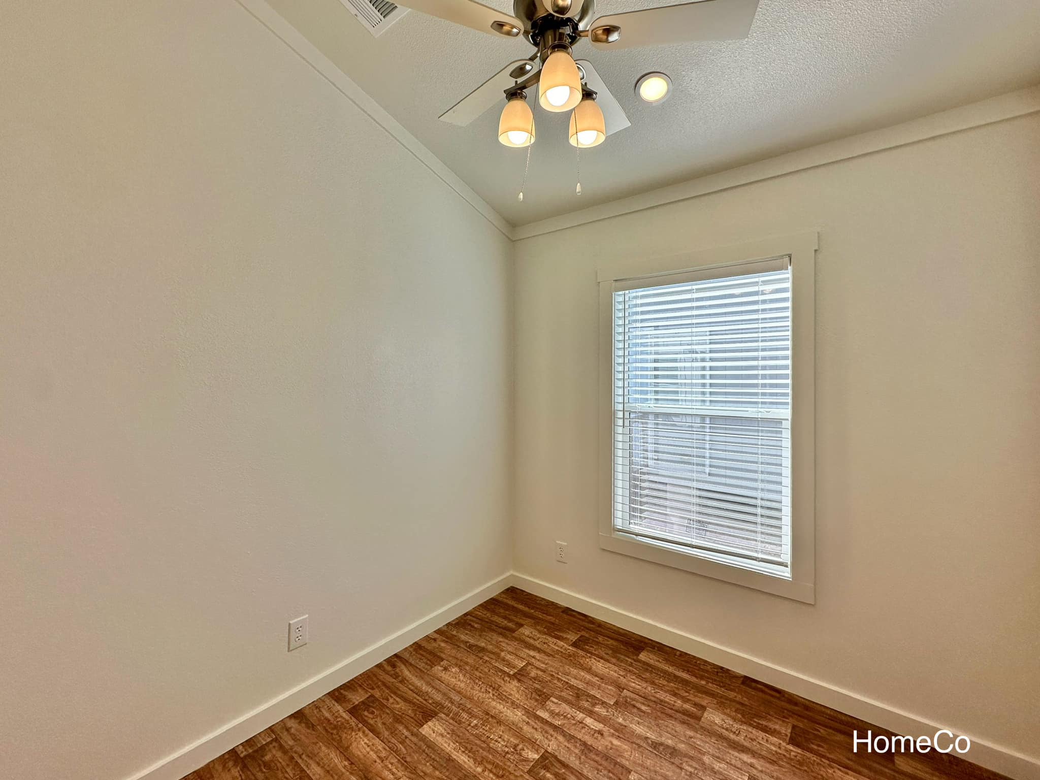 A small, empty room with wooden flooring and cream walls. A white ceiling fan with three lights hangs above. Sunlight filters through a window with blinds.