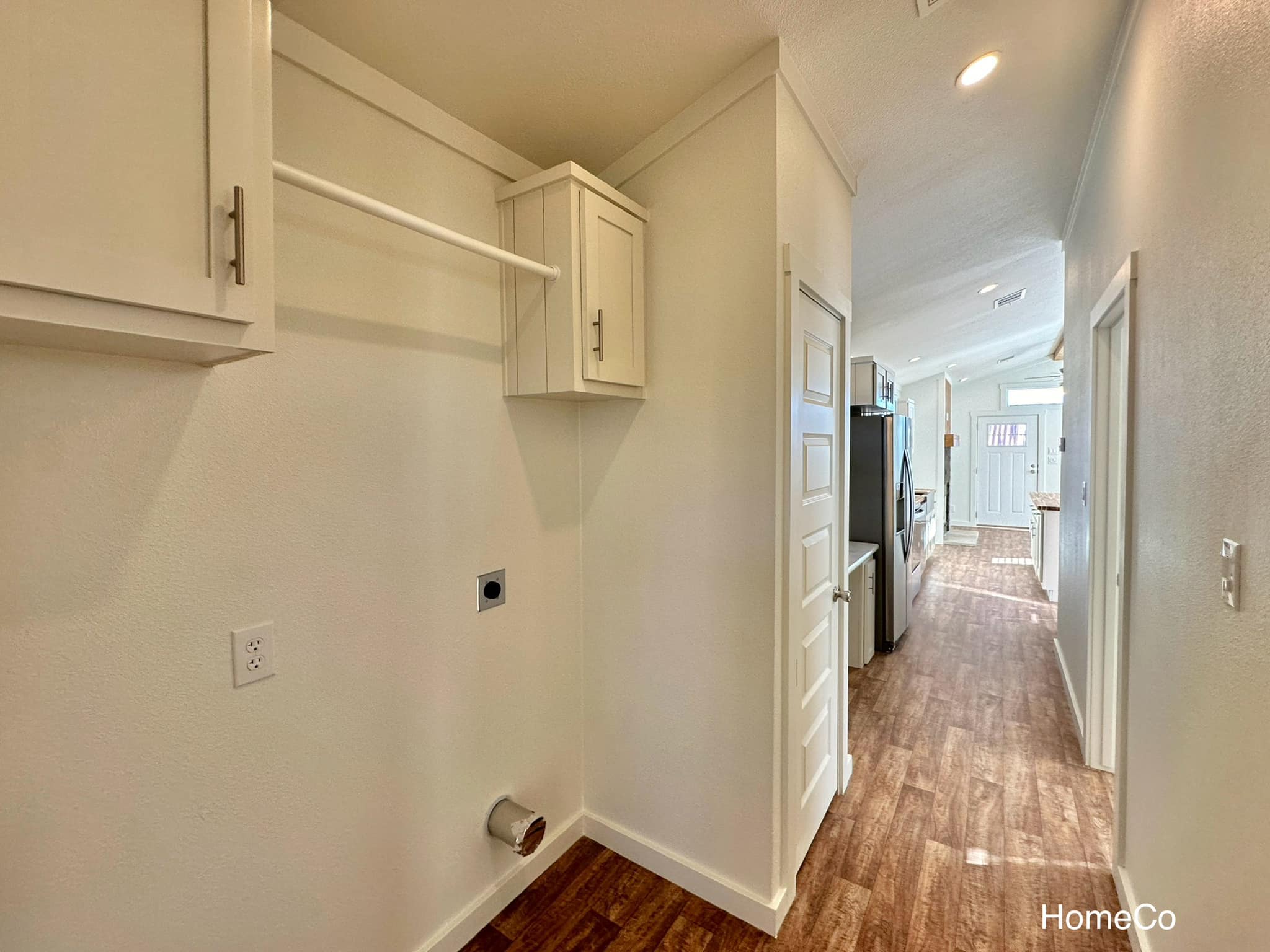 Narrow hallway with wooden floor, white walls, and ceiling lighting. Features a washer-dryer hookup, cabinet, and view into a bright kitchen area.
