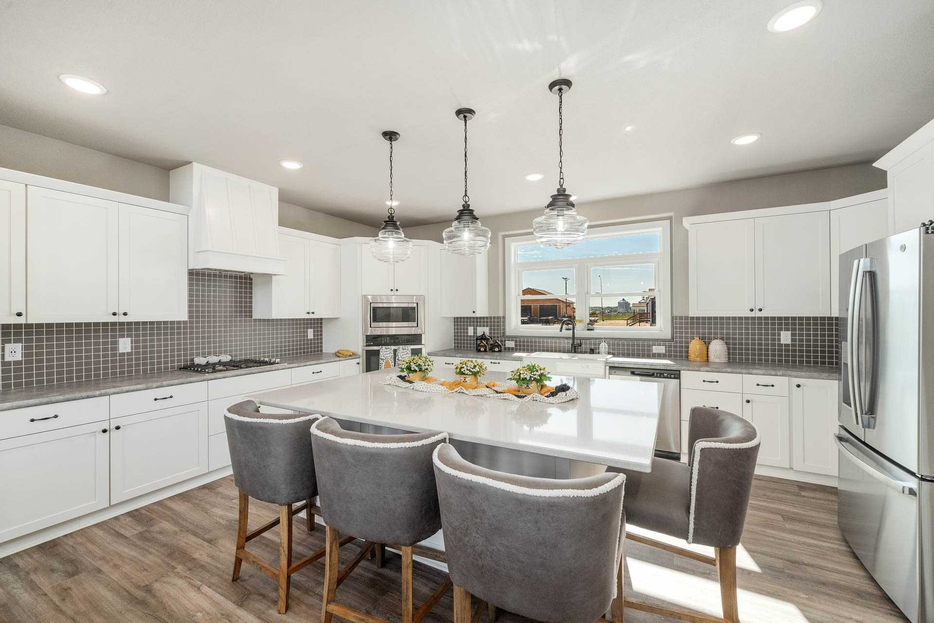 Spacious modern kitchen with white cabinets, large island, and gray chairs. Three pendant lights hang above. Bright and airy with natural light from a window.