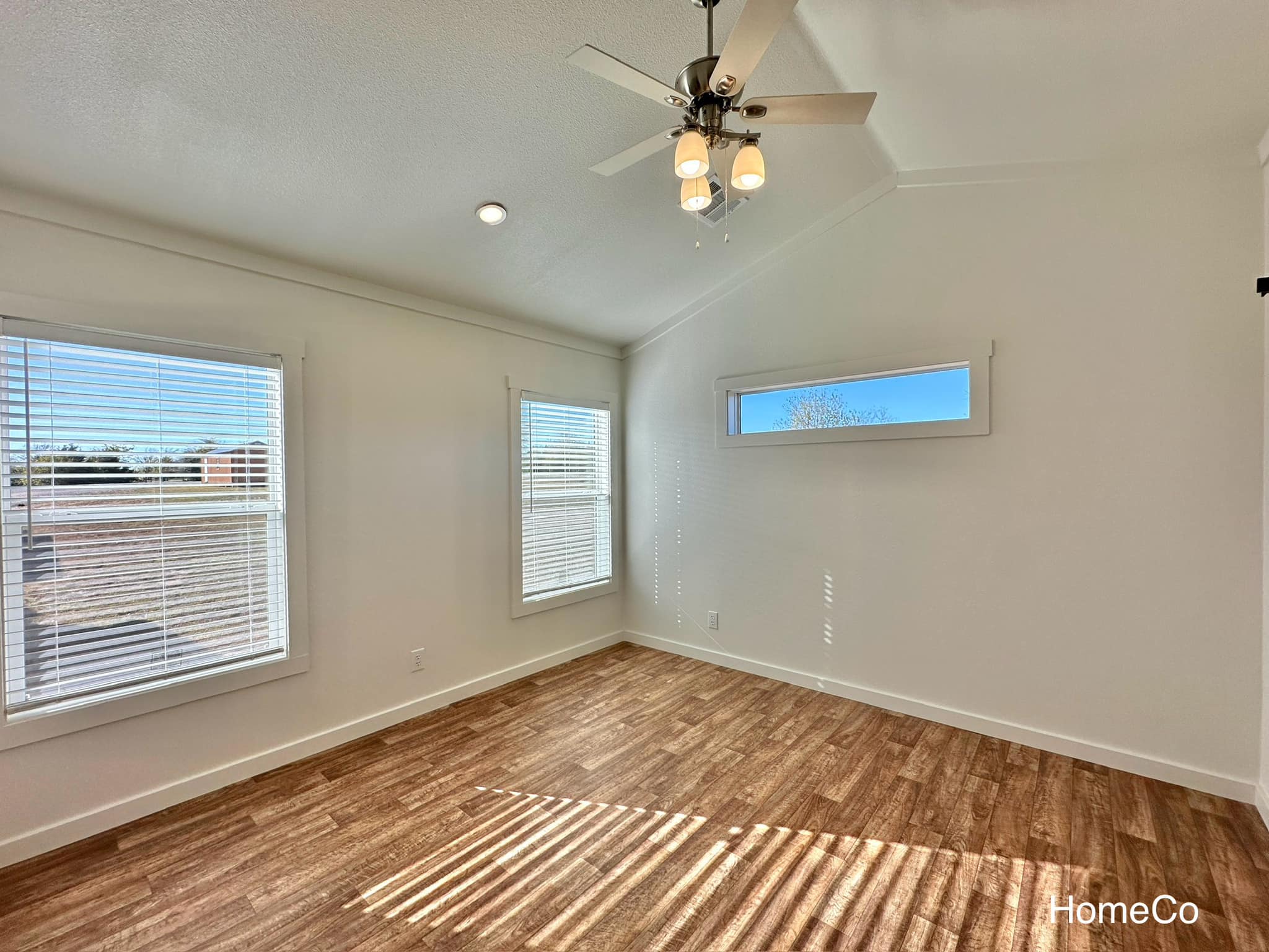 Bright, empty room with wood floors, white walls, and three windows. Ceiling fan and recessed lighting create an airy, inviting ambiance.