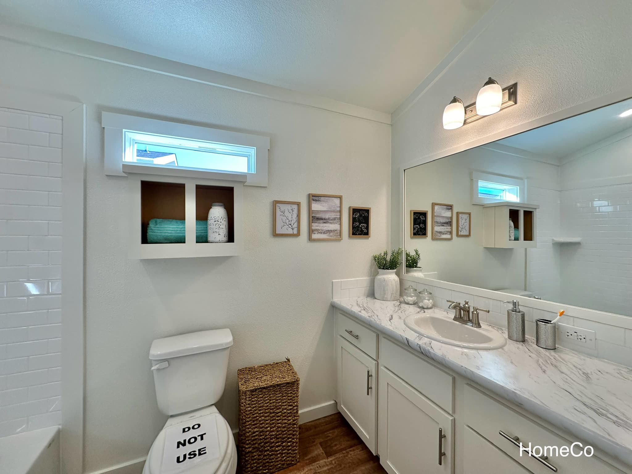 Bright, modern bathroom with white cabinetry and marble countertop. Decor includes wall art, a wicker basket, and a "Do Not Use" cover on the toilet.