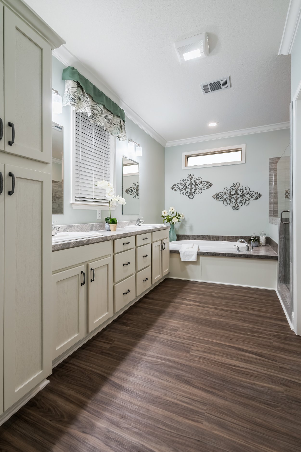 Spacious bathroom with white cabinets, wood floor, and a large mirror. Green valance above the window, elegant metal wall art, and flowers add decor.