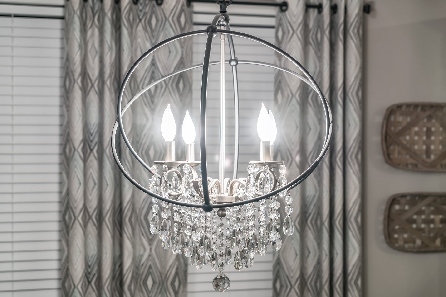 A modern chandelier with four glowing bulbs and hanging crystals inside a spherical metal frame. Geometric curtains and woven baskets adorn the background.