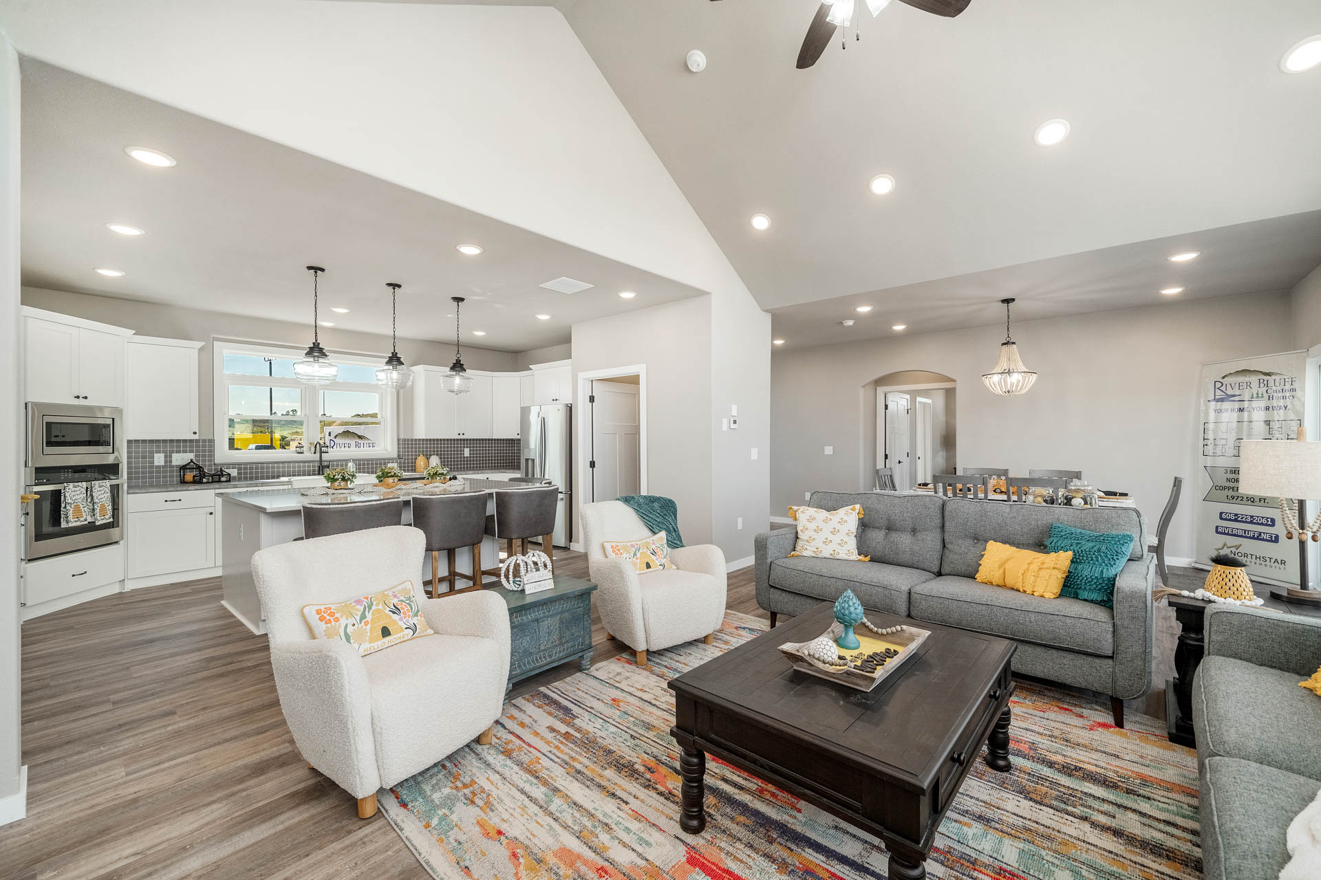 Spacious living room and kitchen with vaulted ceilings. Gray sofas, colorful pillows, and a patterned rug create a cozy, modern ambiance. Bright lighting.
