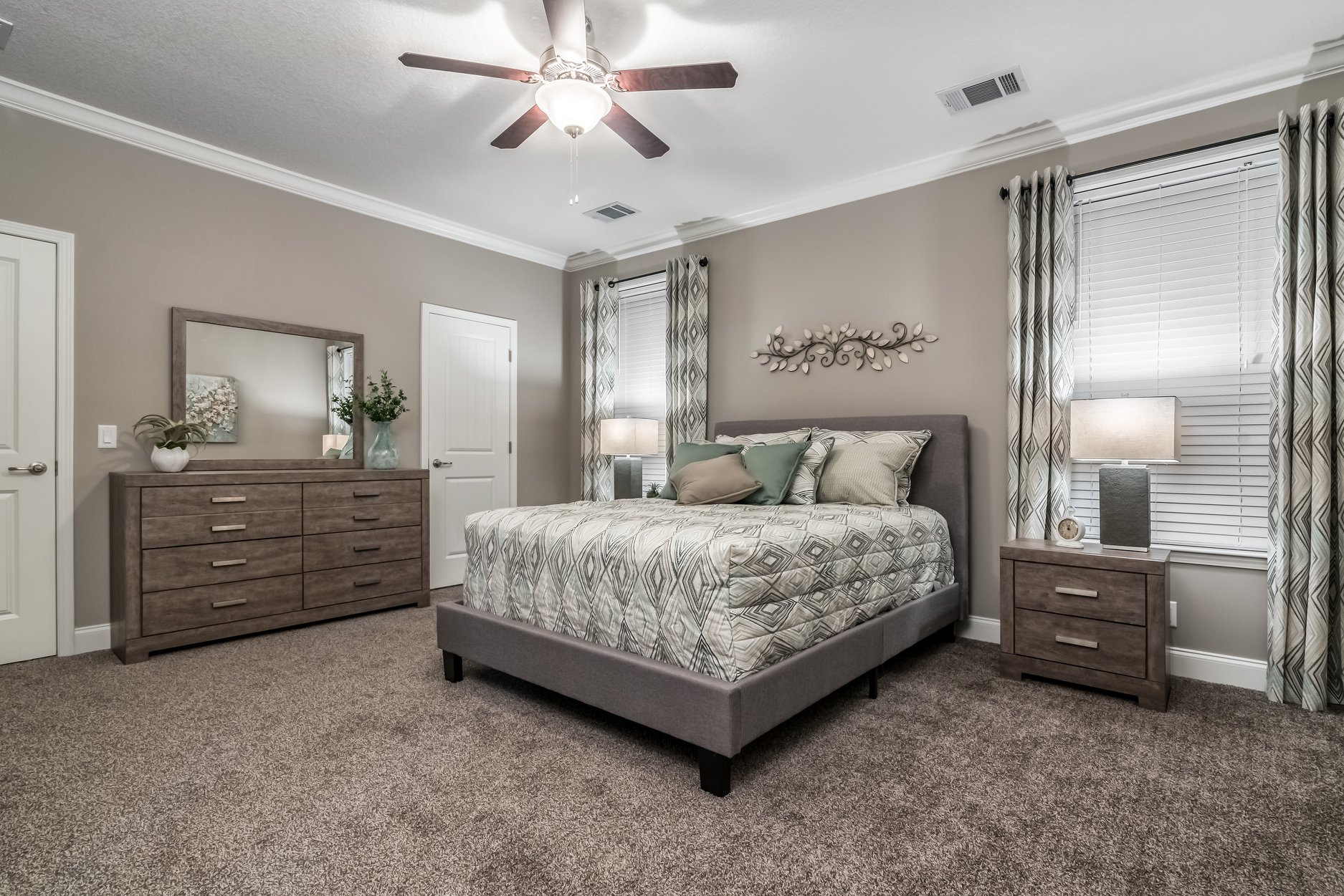 A cozy bedroom with a plush bed featuring patterned bedding and several pillows. Two lamps on dark wood nightstands emit warm light. A ceiling fan is on.
