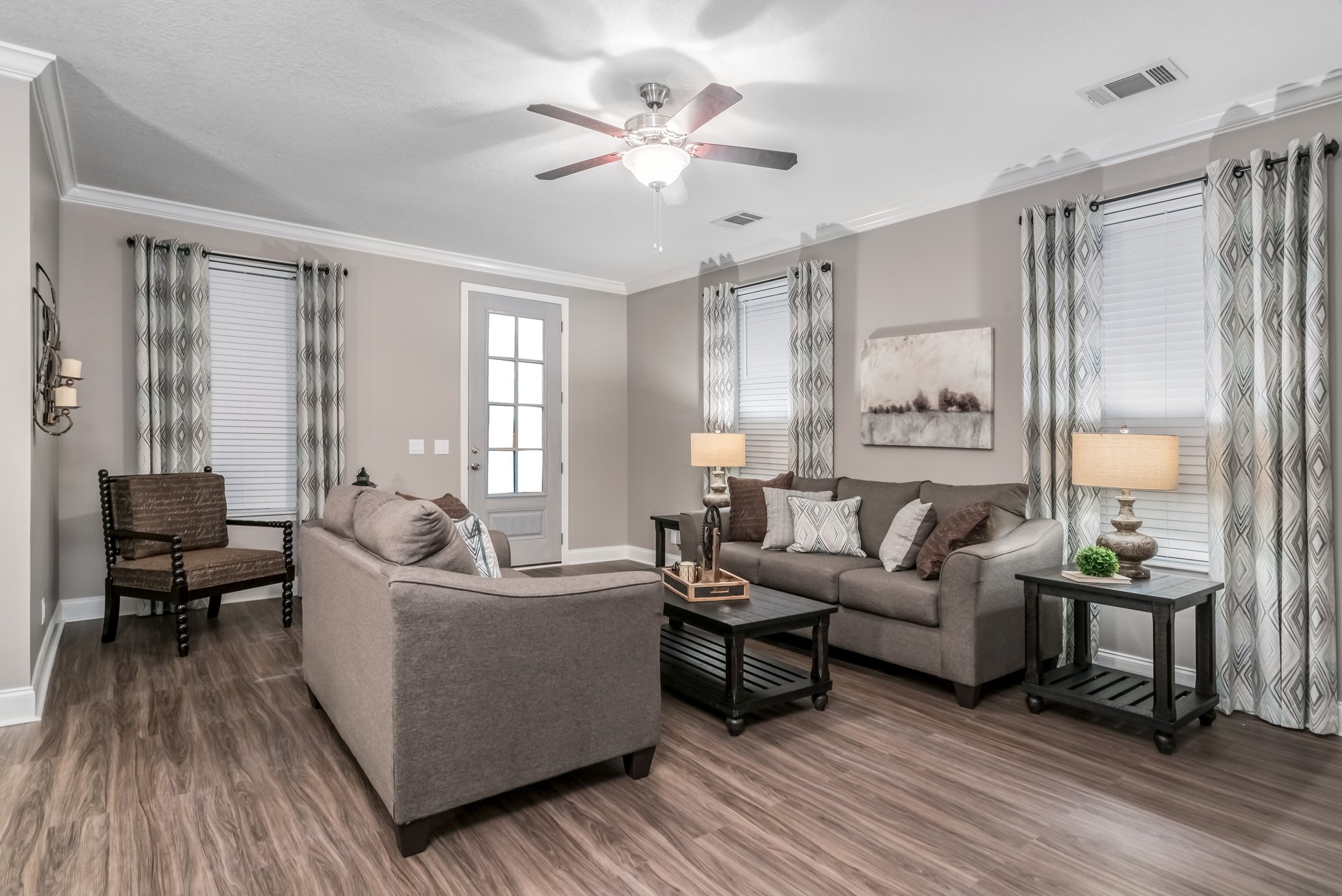 Stylish living room with a gray sofa set, wooden coffee table, and two side lamps. Neutral curtains frame three windows. Ceiling fan overhead. Elegant and inviting atmosphere.