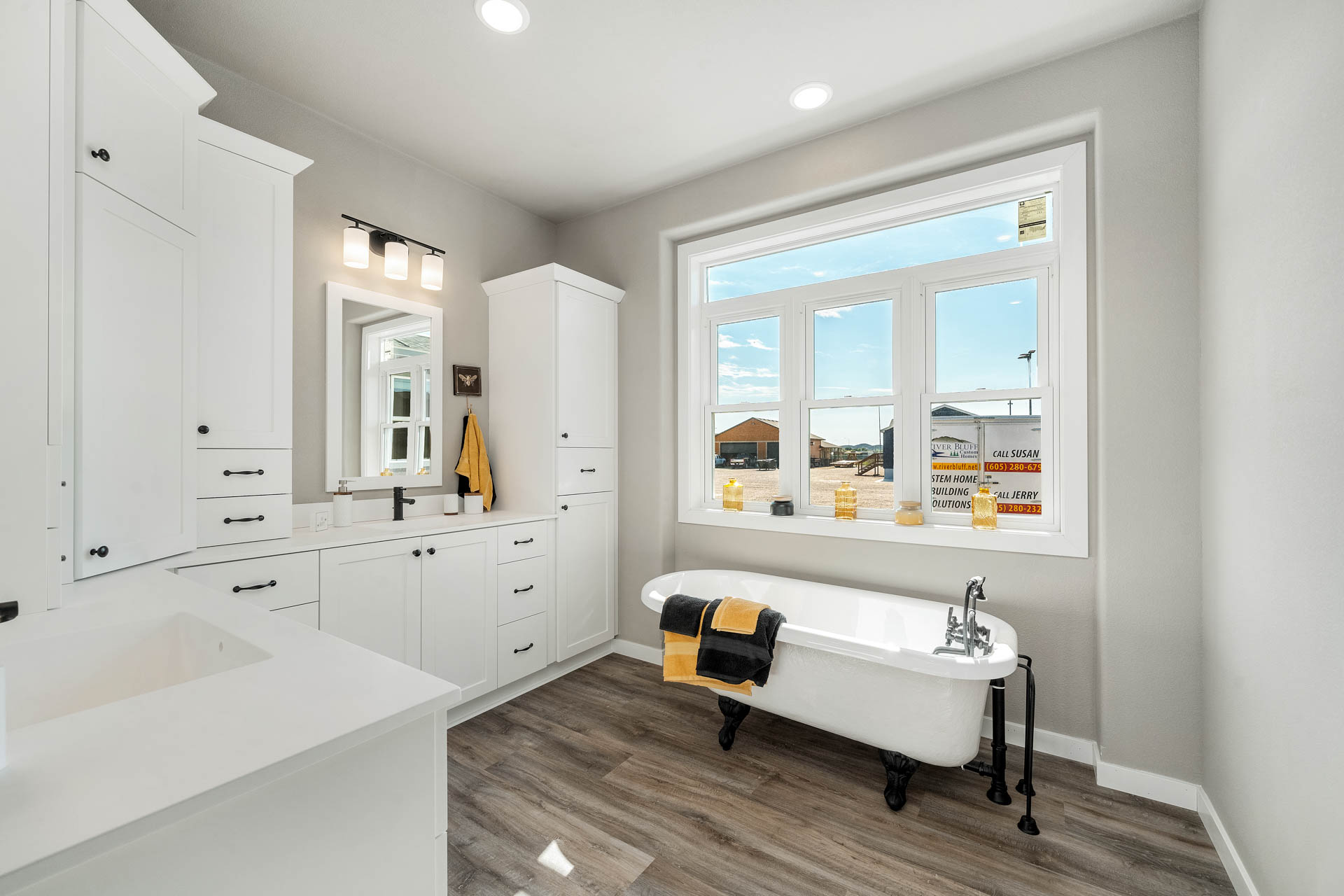 Bright bathroom with a large window, white cabinets, and a vintage clawfoot tub. Yellow and black towels add a cheerful contrast.