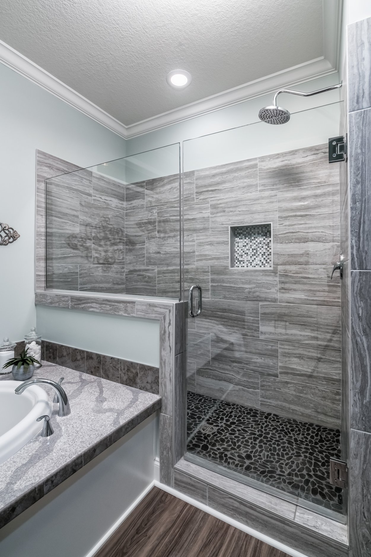 Modern bathroom with a glass-enclosed shower. The walls feature gray tiles and a pebble stone floor. A white bathtub and wood floor add warmth.