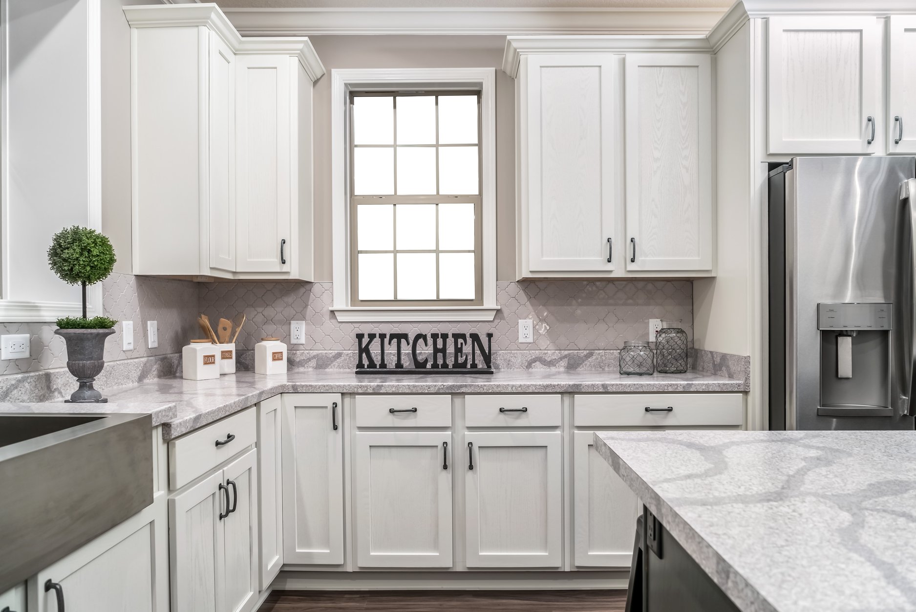 Bright, modern kitchen with white cabinets, marble countertops, and a stainless steel fridge. A window and "KITCHEN" sign add charm. Cozy and inviting.