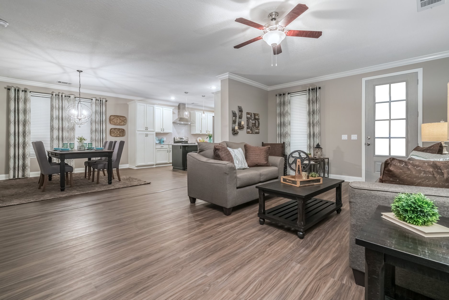 Modern open-concept living room with neutral tones. Gray sofa and brown cushions face a black coffee table. Dining area and kitchen are visible, creating a cozy, welcoming atmosphere.