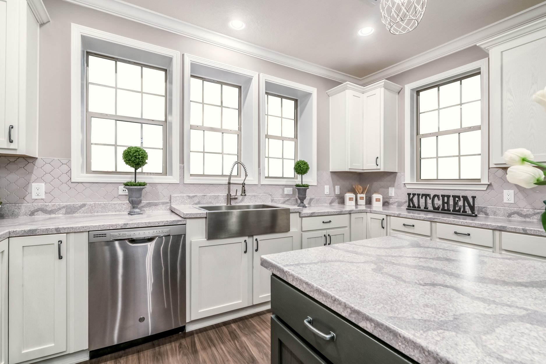 Bright kitchen with white cabinets and grey countertops, featuring a farmhouse sink, large windows, stainless steel appliances, and decorative plants.