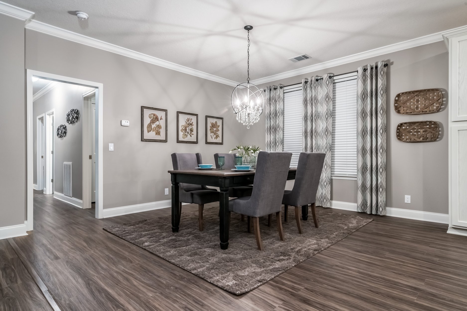 Elegant dining room with a modern chandelier, gray upholstered chairs, and a black table on a brown rug. Artwork and wicker decor adorn light walls.