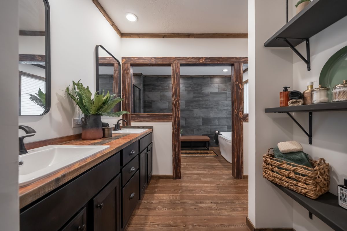 Rustic modern bathroom with dark wood accents, dual sinks, and a large mirror. Gray tile shower, freestanding tub, and open shelves evoke elegance.