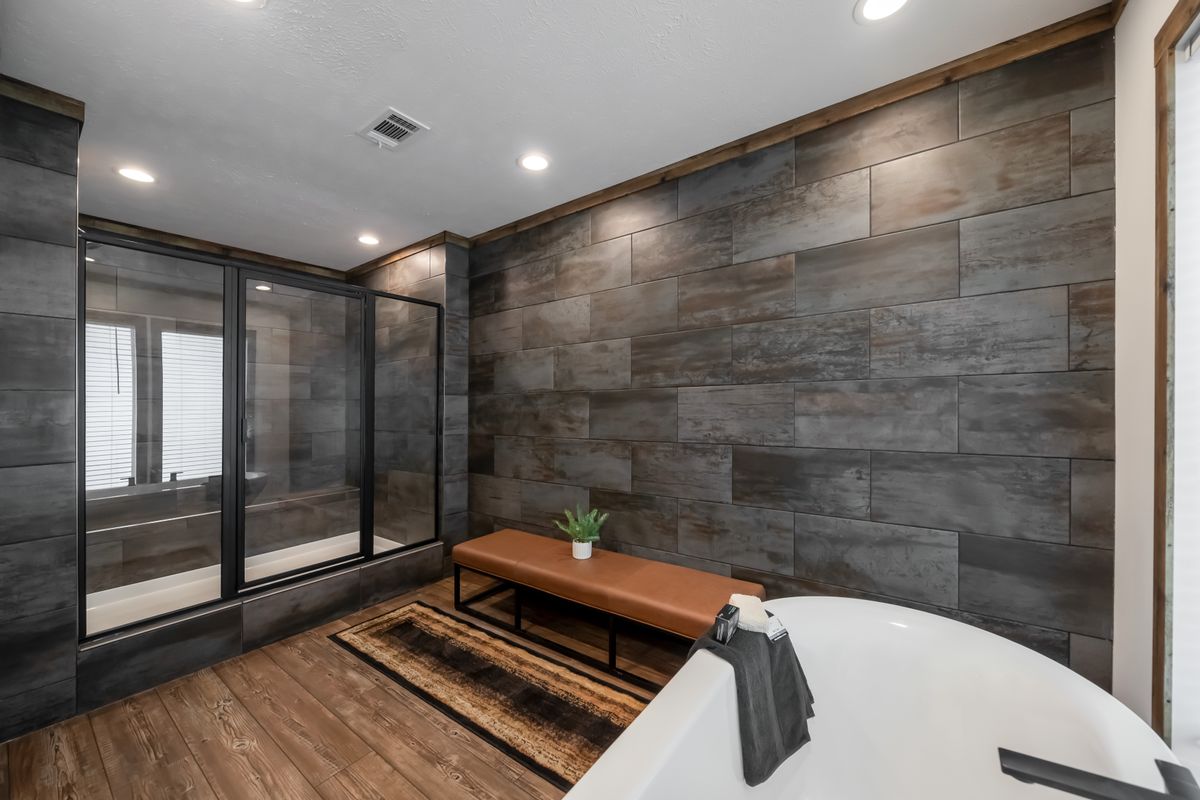 Modern bathroom with sleek gray tiles and wood elements. A brown bench with a plant adds warmth. Large glass shower and white tub create a calm ambiance.
