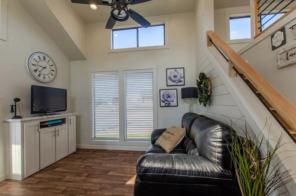 Modern living room with a black leather sofa, wall clock, and TV on a white stand. Bright natural light filters through dual blinds; a cozy, inviting space.