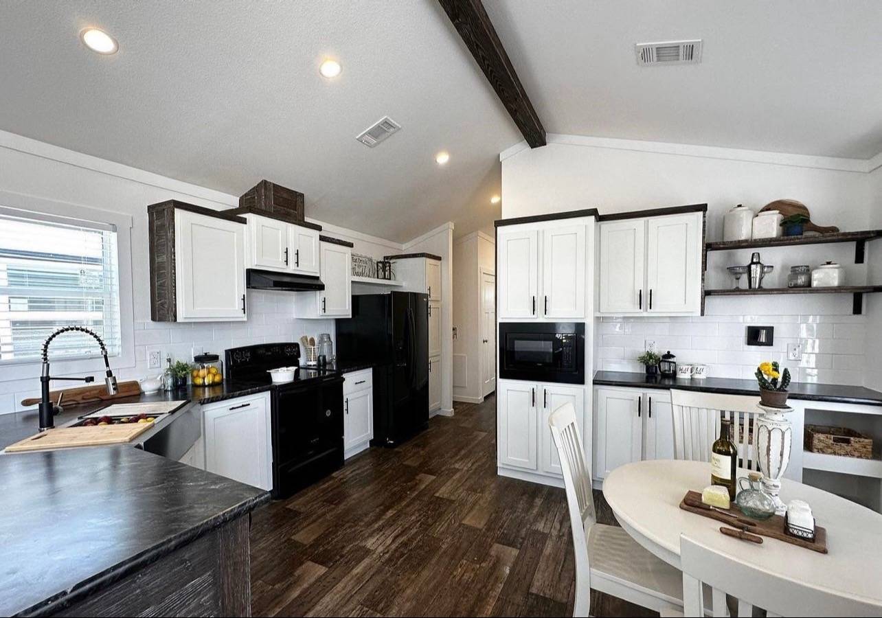 Modern kitchen with dark wood floors and white cabinets. Black appliances, farmhouse sink, round dining table with wine, and decorative shelves enhance the cozy ambiance.