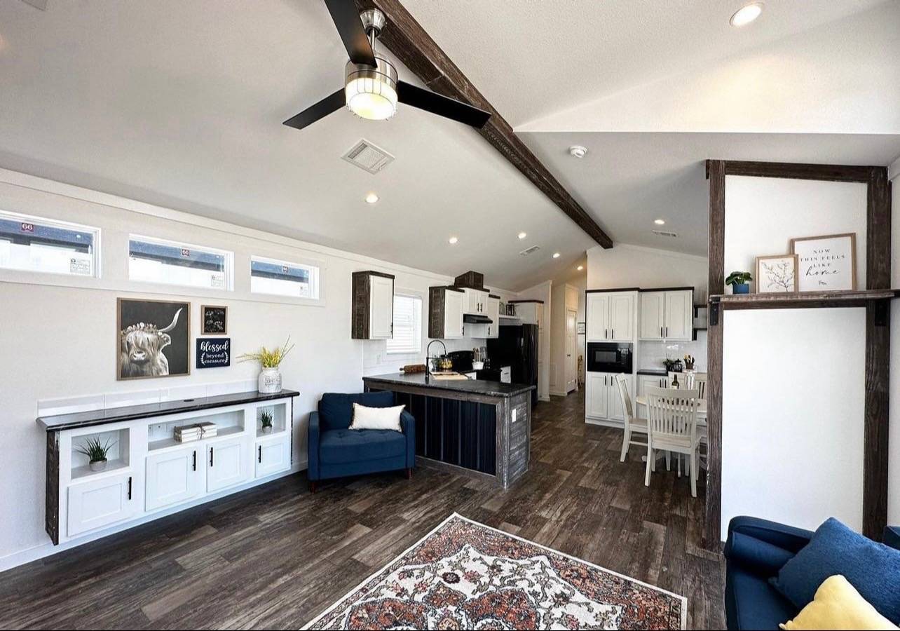 Modern living room and kitchen with wooden floors, exposed beams, and white walls. Blue armchairs, white cabinets, and a central island add elegance.