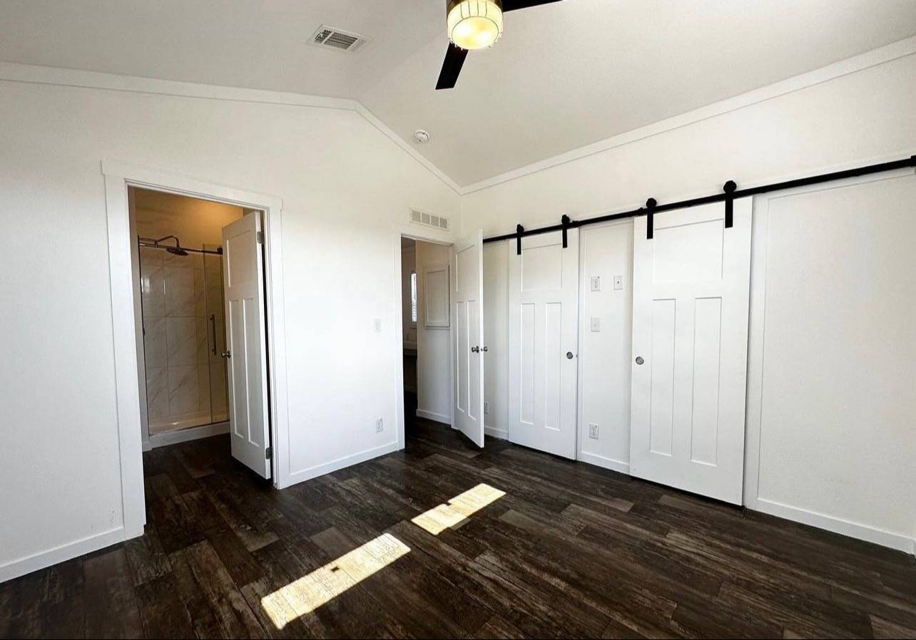 Modern bedroom with wood floors, white walls, and a ceiling fan with light. Features sliding barn doors and an open door to a bathroom with a shower.