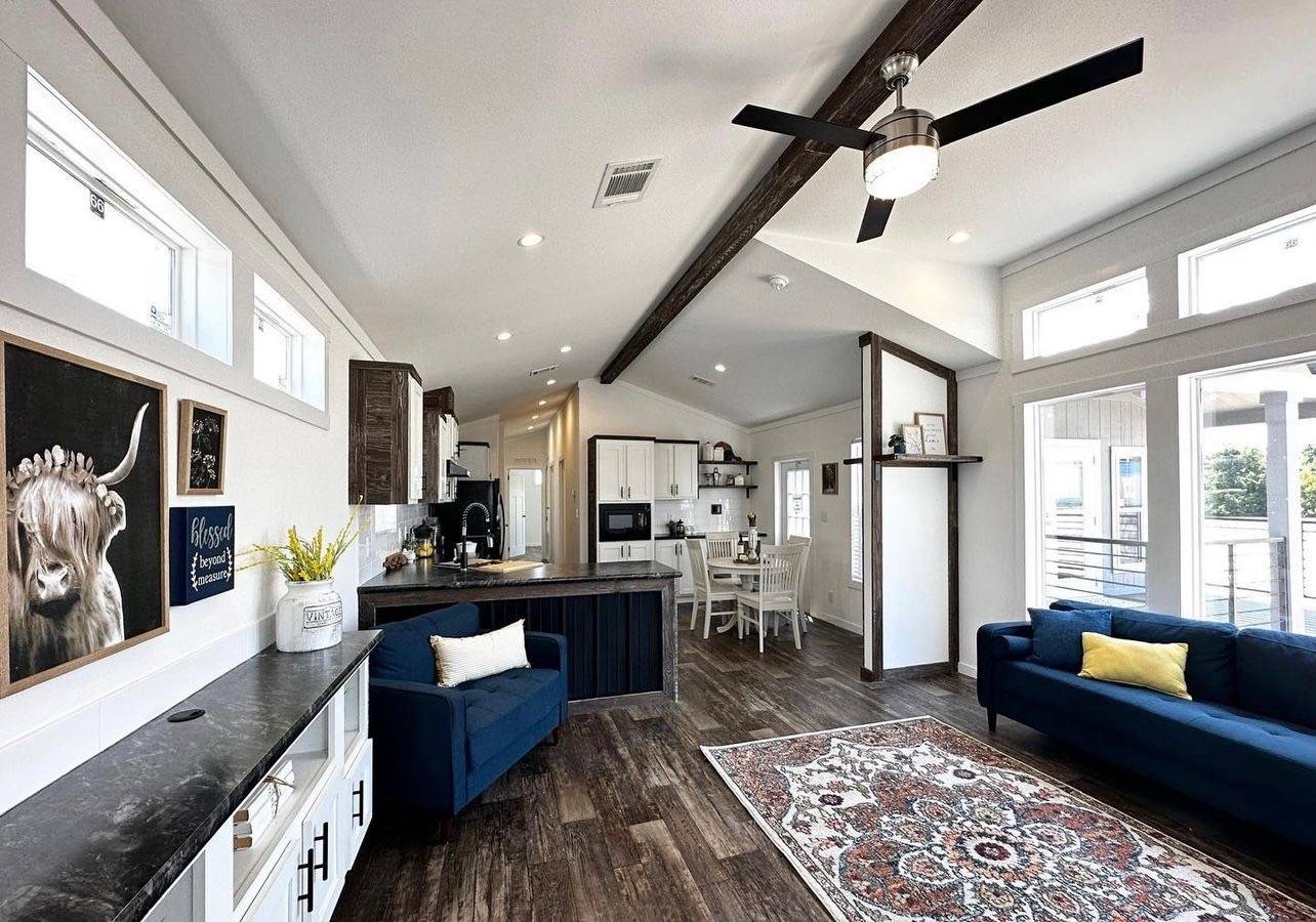 Spacious modern living room and kitchen with vaulted ceilings, dark wood floors, and large windows. Features blue sofas, a patterned rug, and wall art.