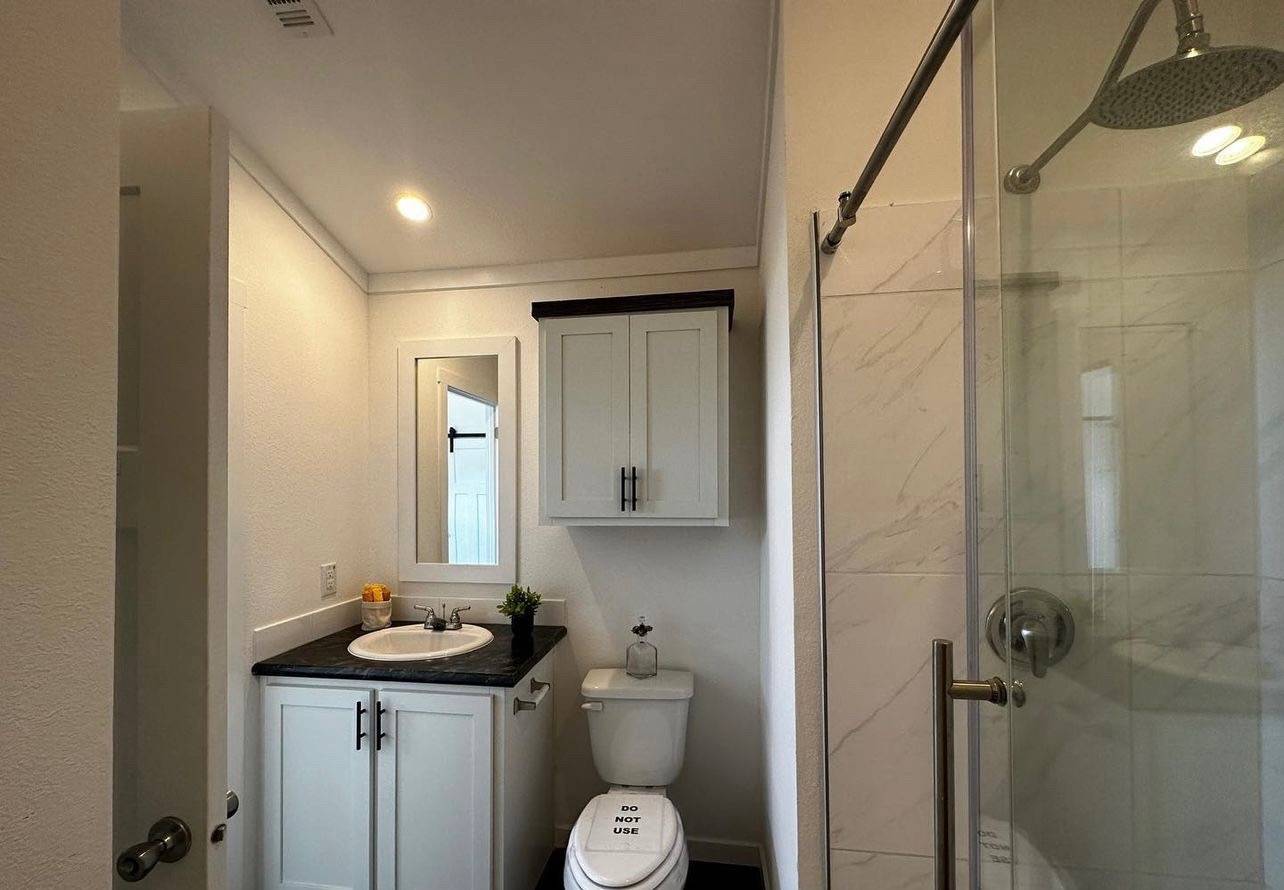 Compact bathroom with a modern shower, white vanity, mirror, and cabinet. A toilet with a "do not use" sign. Bright, clean ambiance.