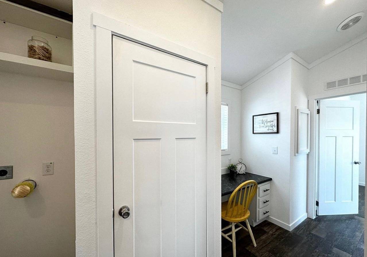 A cozy room features a white door, a small desk with a black countertop, a yellow chair, framed wall art, and a small potted plant, creating a warm, inviting space.
