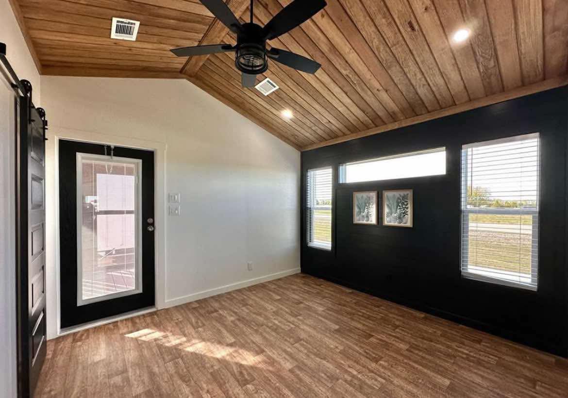 Modern room with wood ceiling and floor, black accent wall, and large windows. A ceiling fan hangs overhead, creating a cozy, inviting atmosphere.