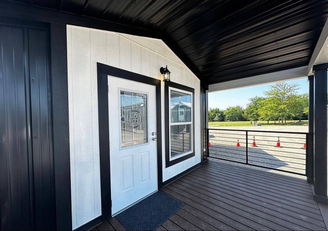 Small house entrance with a white door featuring a decorative glass star. Black trim and siding are prominent. Porch overlooks a gravel area with traffic cones and trees in the background.